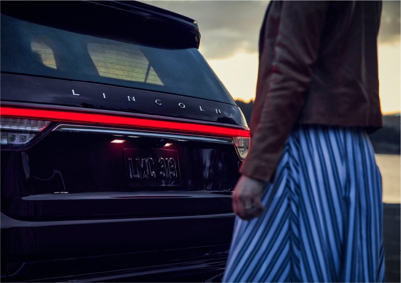 A person is shown near the rear of a 2023 Lincoln Aviator® SUV as the Lincoln Embrace illuminates the rear lights | Dave Sinclair Lincoln in St Louis MO